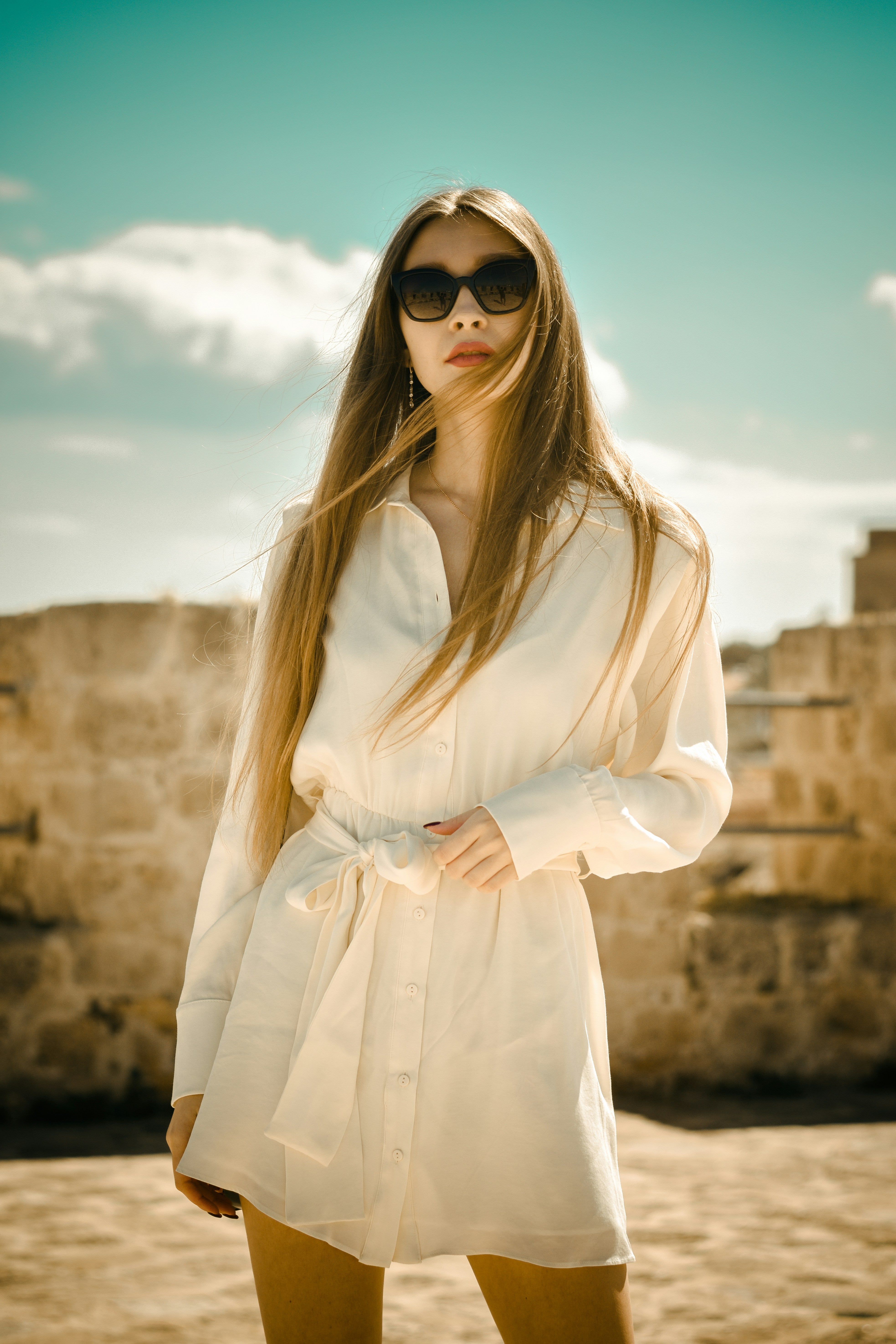 Woman wearing a white shirt and sunglasses with a stone wall and blue sky in the background