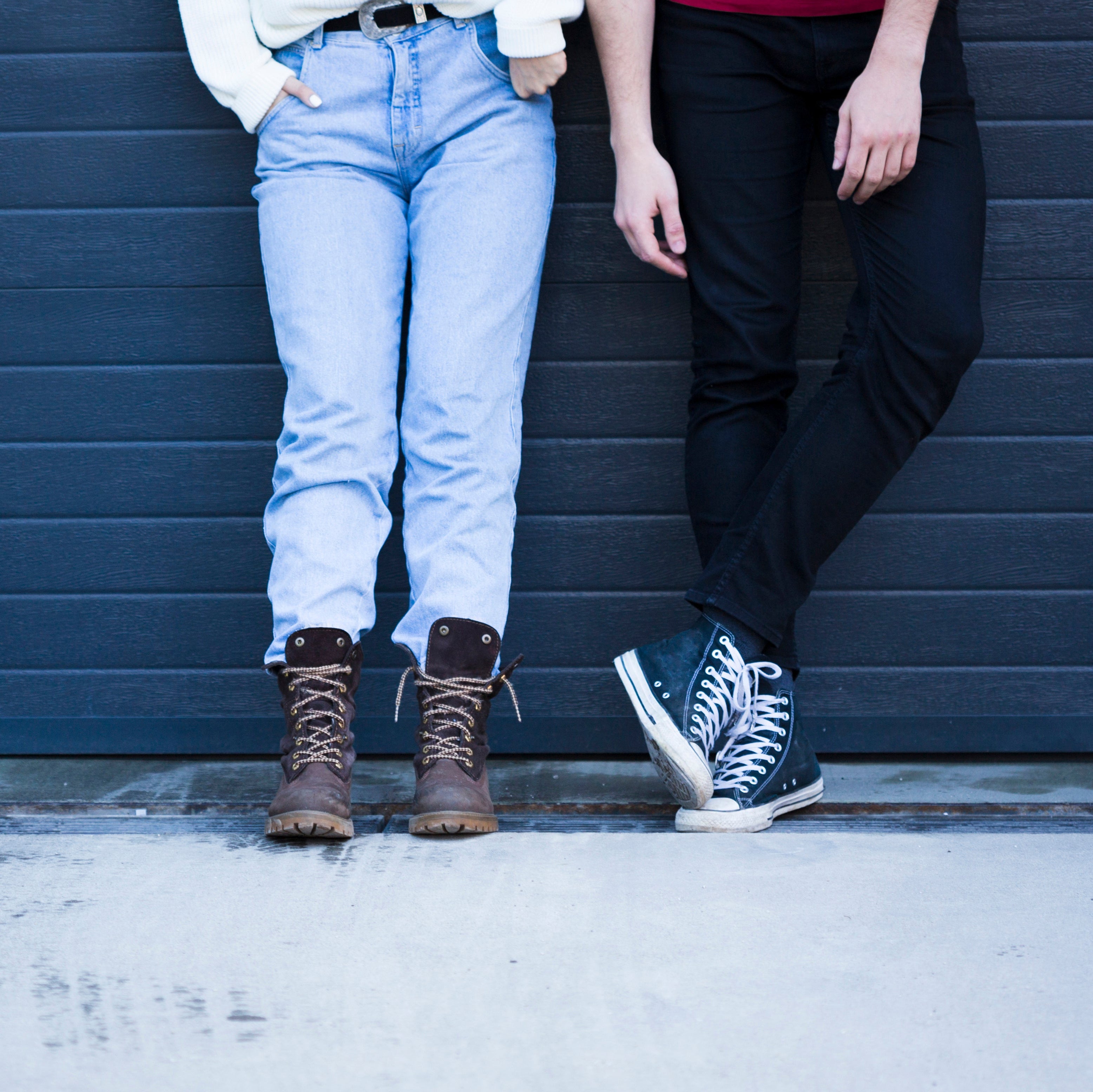 Two people standing against a dark blue wall, wearing jeans and boots.